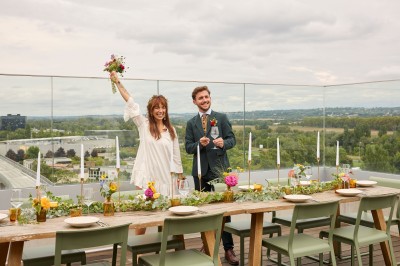 Un couple célèbre son mariage sur une petite scène décorée de voiles et de feuillages, devant une fresque colorée.