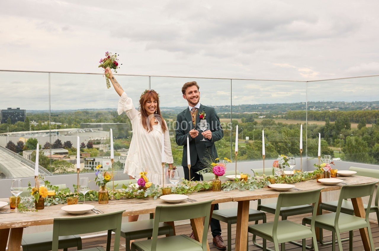 Un couple souriant debout sur une terrasse décorée, tenant des fleurs et des verres, avec une vue panoramique en arrière…