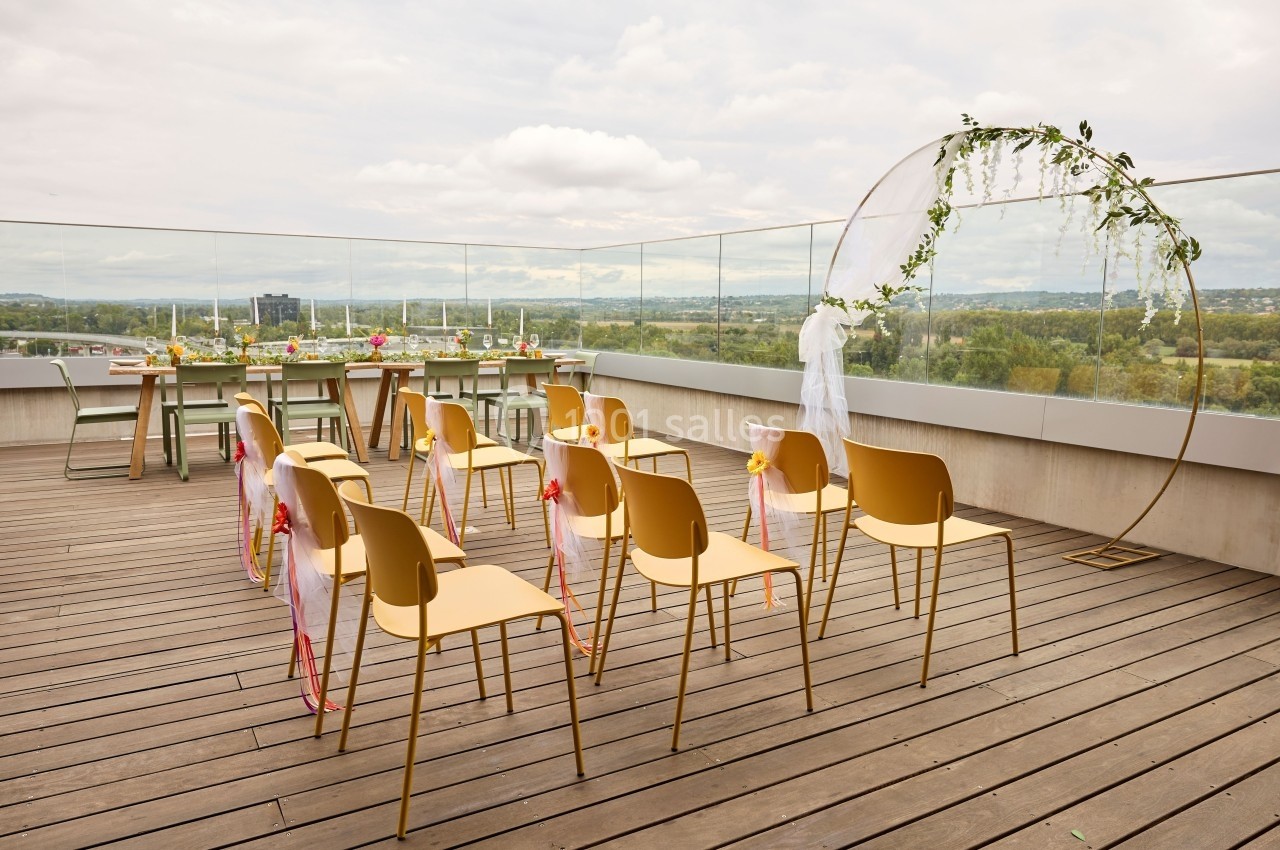 Chaises alignées sur une terrasse en bois avec arche décorée et vue dégagée sur un paysage verdoyant.