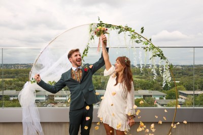 Un couple célèbre son mariage sur une petite scène décorée de voiles et de feuillages, devant une fresque colorée.