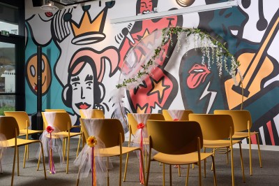 Un couple célèbre son mariage sur une petite scène décorée de voiles et de feuillages, devant une fresque colorée.