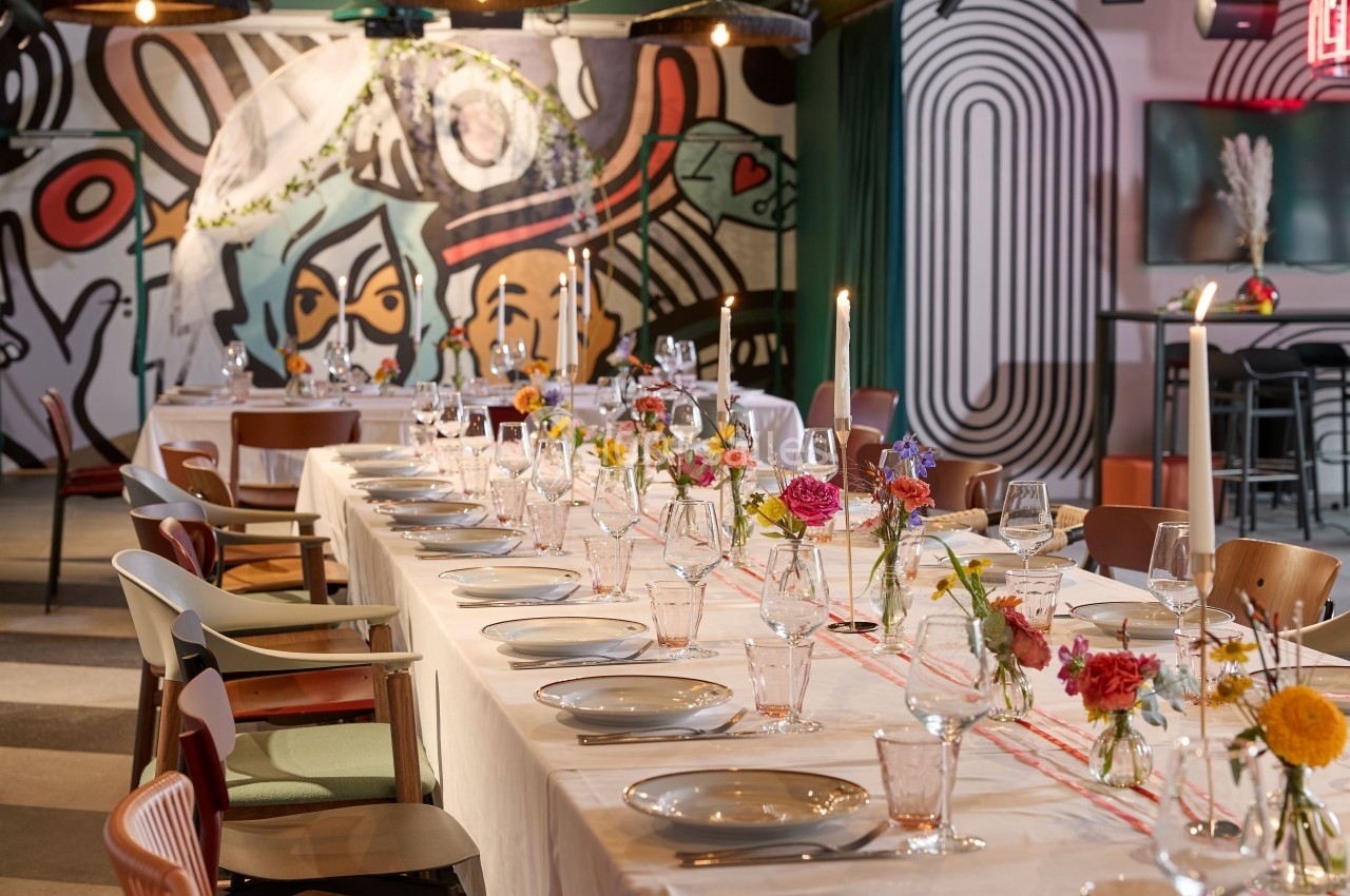 Table dressée avec vaisselle et bougies, décorée de fleurs colorées, dans une salle au mur orné de fresques modernes.