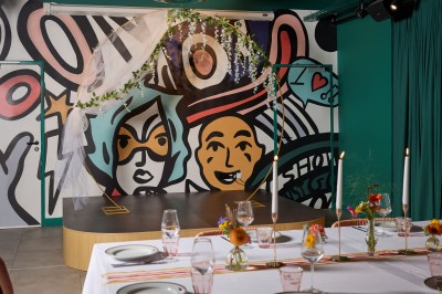 Un couple célèbre son mariage sur une petite scène décorée de voiles et de feuillages, devant une fresque colorée.