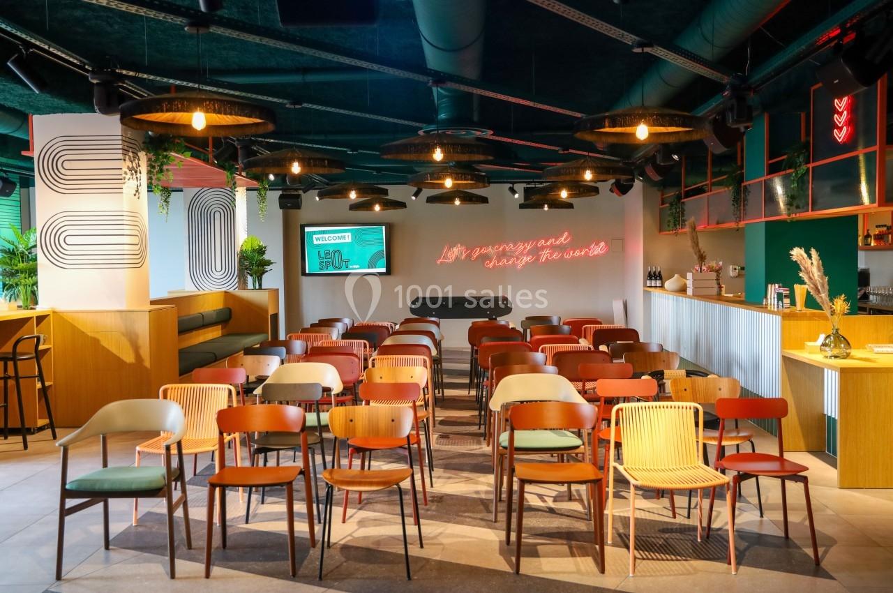 Salle lumineuse aménagée pour une présentation, avec des rangées de chaises colorées et un écran au fond.