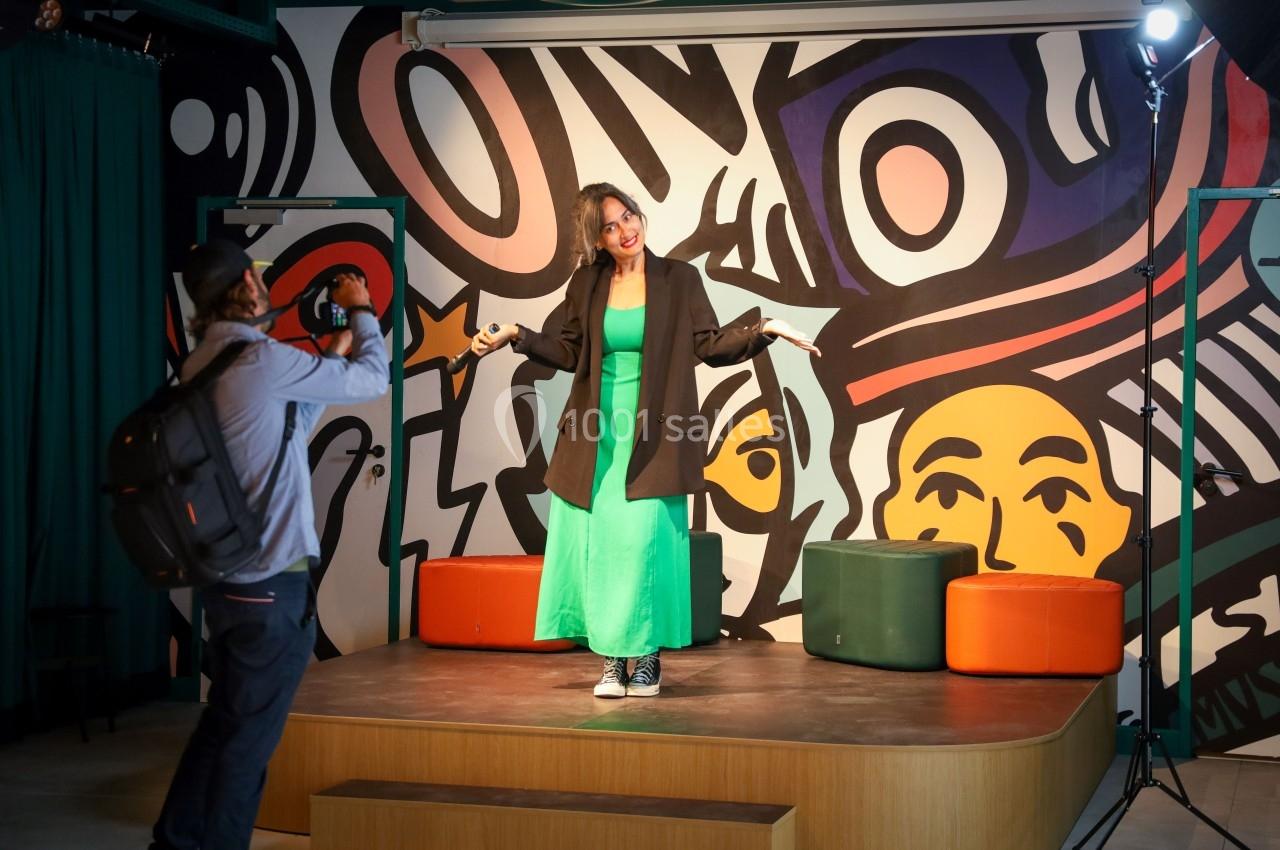 Une femme pose sur une petite scène devant une fresque colorée, photographiée par une personne avec un appareil photo.