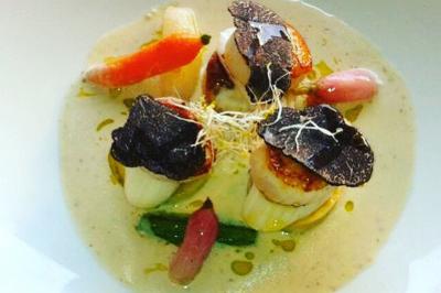 Homme en veste de chef blanche, debout devant une architecture en pierre et des grilles métalliques.