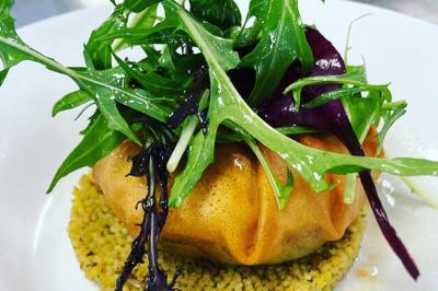 Homme en veste de chef blanche, debout devant une architecture en pierre et des grilles métalliques.