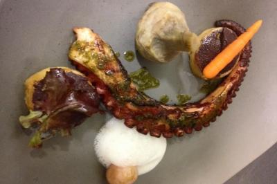 Homme en veste de chef blanche, debout devant une architecture en pierre et des grilles métalliques.