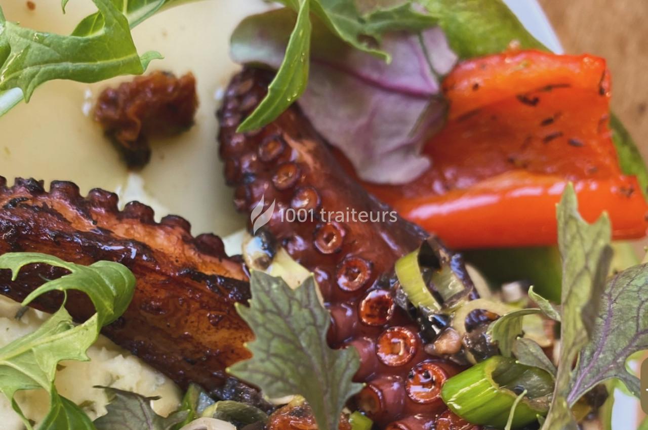 Tentacule de poulpe grillé accompagné de légumes colorés, herbes fraîches et fromage sur une assiette.
