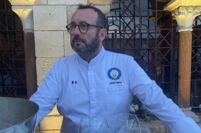 Homme en veste de chef blanche, debout devant une architecture en pierre et des grilles métalliques.