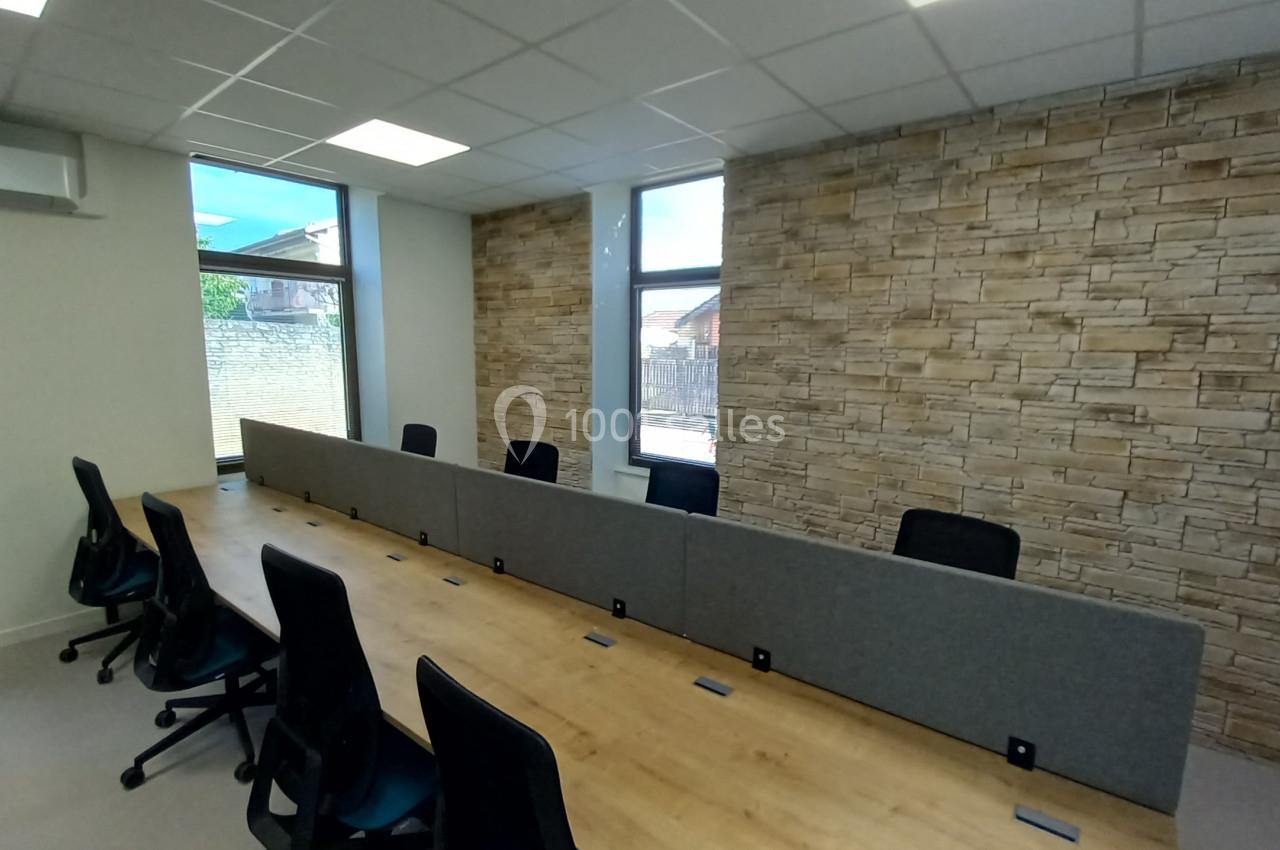 Salle de réunion avec une grande table en bois, chaises noires, cloison centrale et mur en pierre apparente.