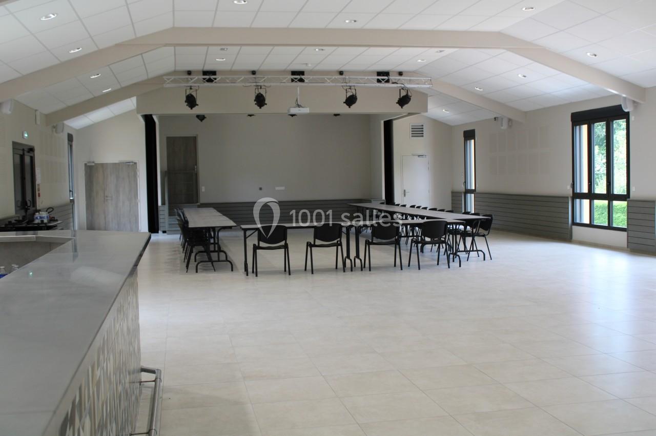 Salle polyvalente lumineuse avec tables et chaises disposées, grandes fenêtres et éclairage au plafond.