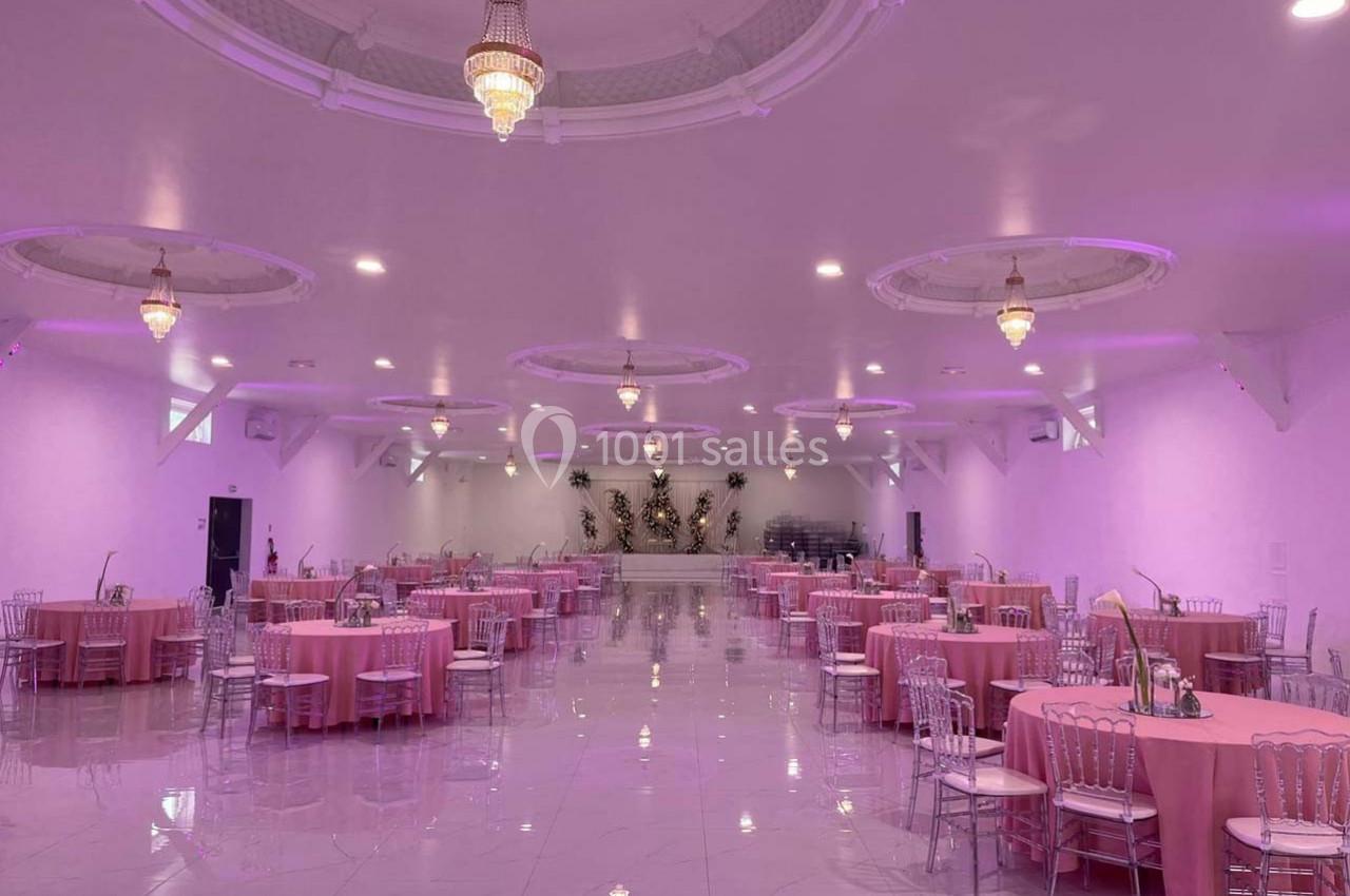 Salle de réception spacieuse avec tables rondes dressées de nappes roses et lustres suspendus au plafond.