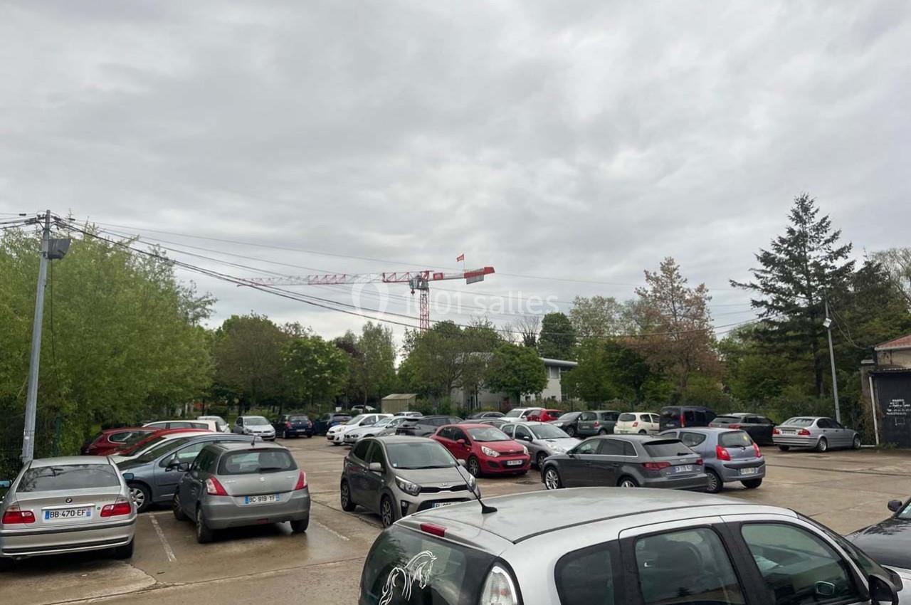 Parking extérieur avec plusieurs voitures stationnées, entouré d'arbres, sous un ciel nuageux, avec une grue visible au loin.
