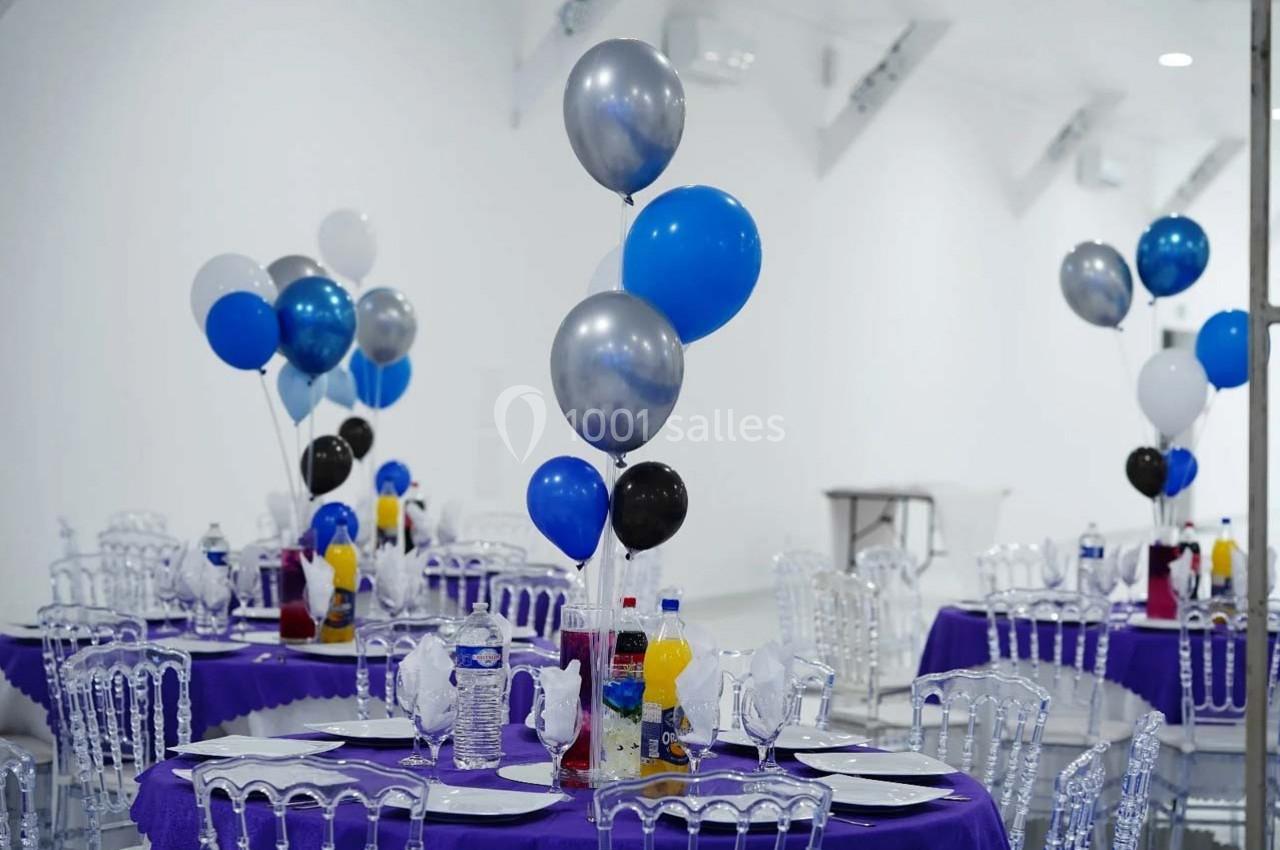 Tables décorées avec nappes violettes, ballons colorés et chaises transparentes dans une salle lumineuse.