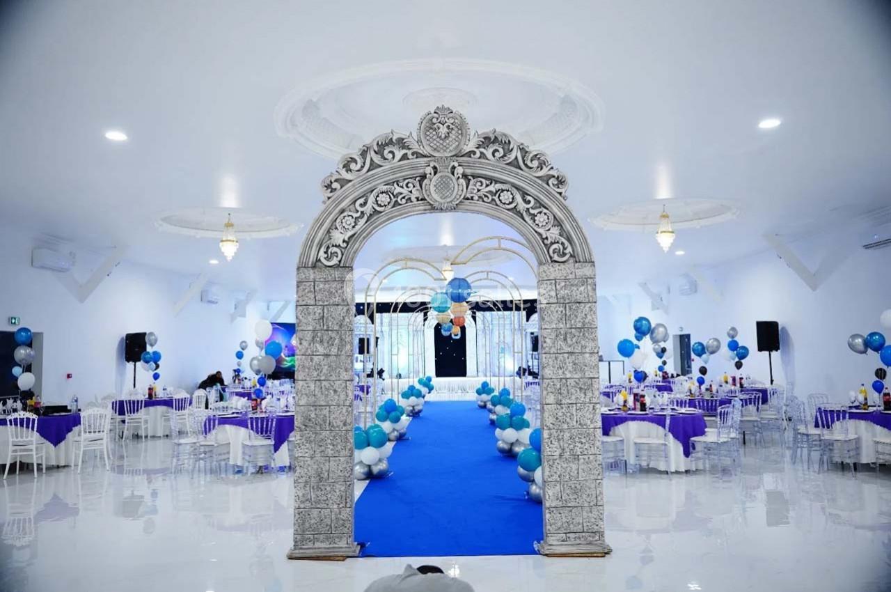 Salle de réception décorée avec des ballons bleus et blancs, tables dressées et arche ornée en premier plan.