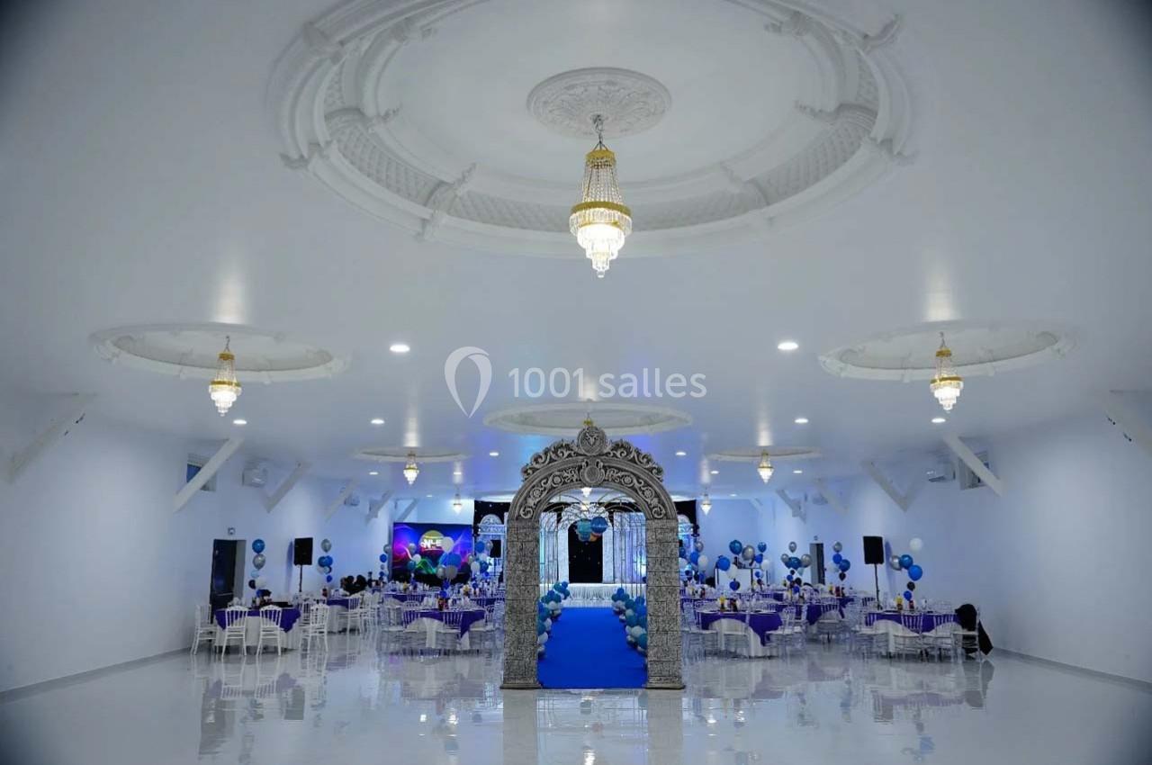 Salle de réception décorée avec des tables dressées, des ballons bleus et une arche ornée au centre.