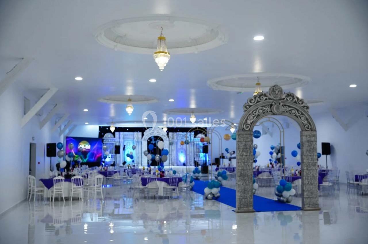 Salle de réception décorée avec des arches, des ballons bleus et blancs, et des tables dressées pour un événement.