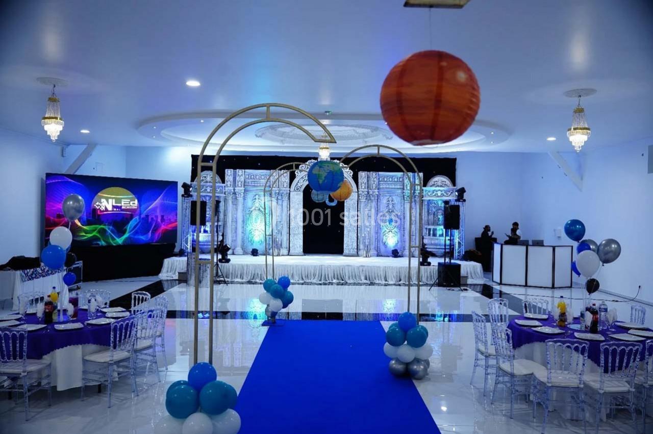 Salle de réception décorée avec des ballons, une scène illuminée et des tables dressées pour un événement festif.