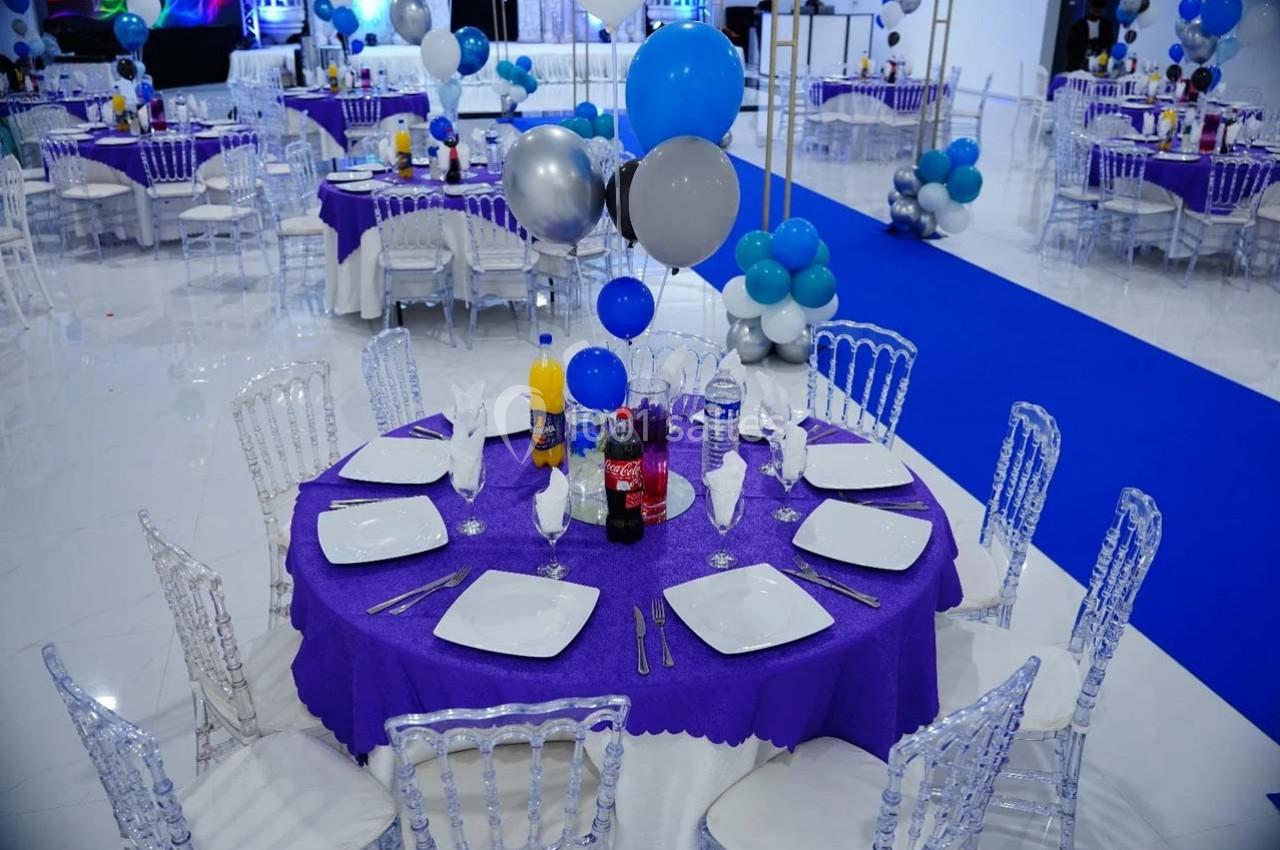 Salle décorée pour un événement avec tables rondes violettes, chaises transparentes et ballons bleus, argentés et blancs.