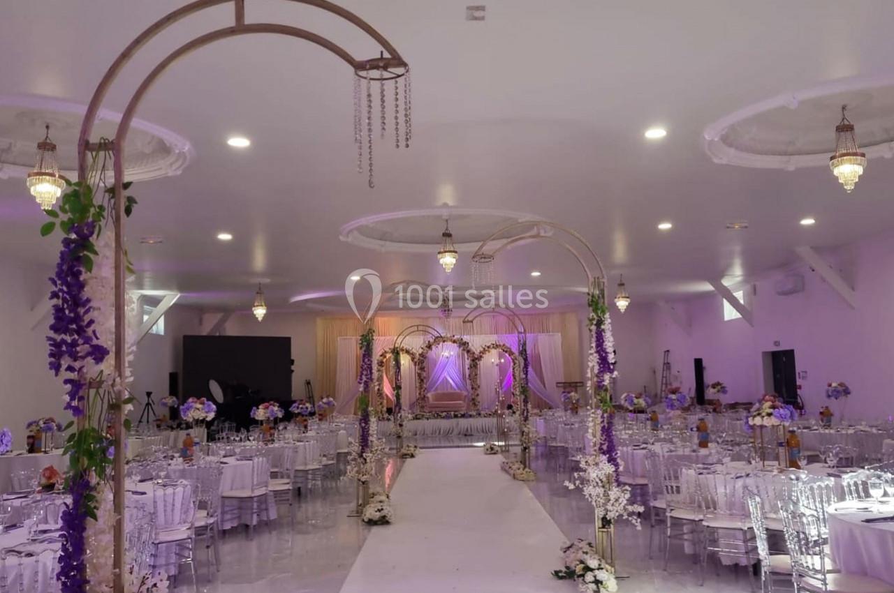 Salle de réception décorée avec des arches fleuries, des tables dressées et un éclairage violet.