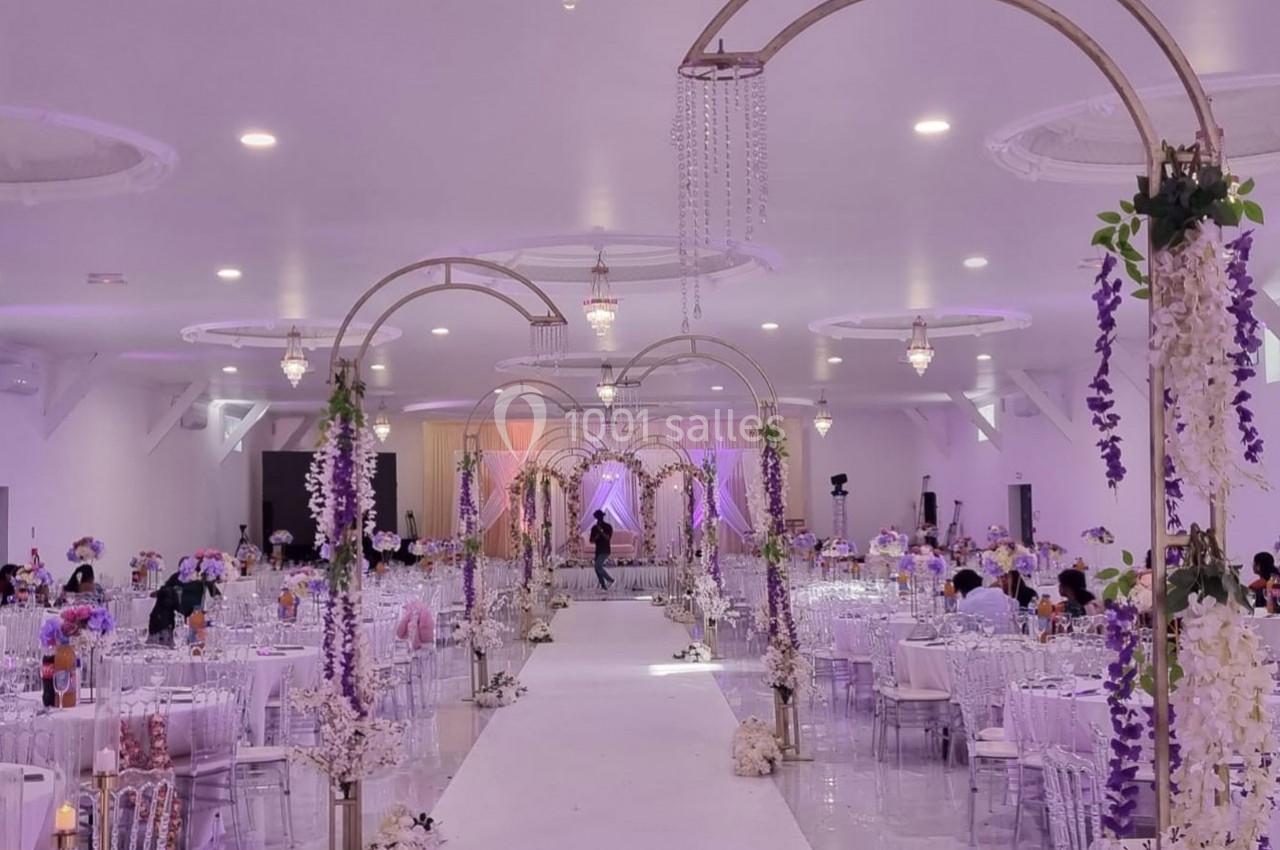 Salle de réception décorée avec des arches fleuries, des tables dressées et un éclairage doux dans des tons violets.