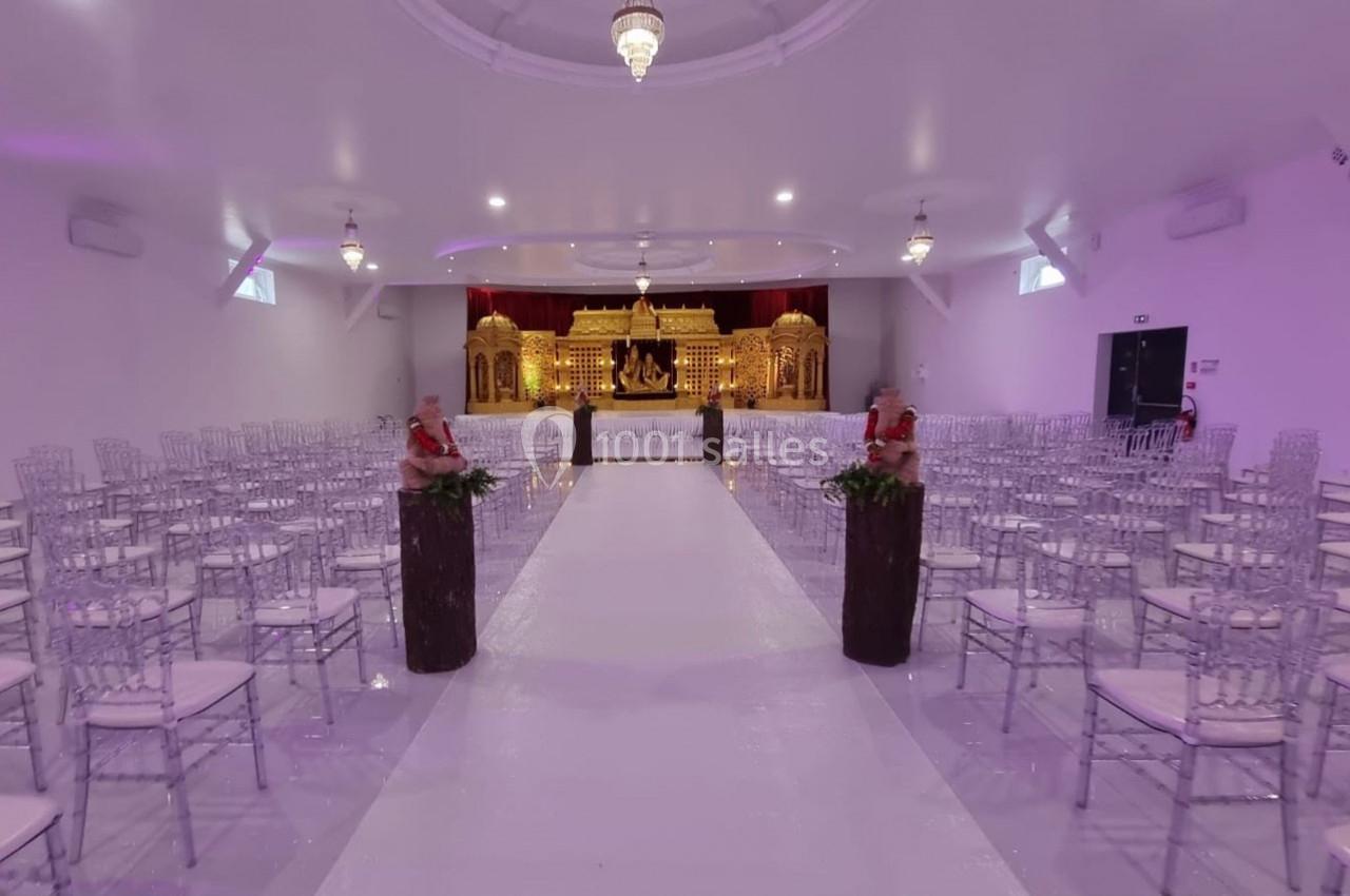 Salle de réception décorée avec des chaises transparentes, une allée centrale blanche et une scène ornée de dorures.
