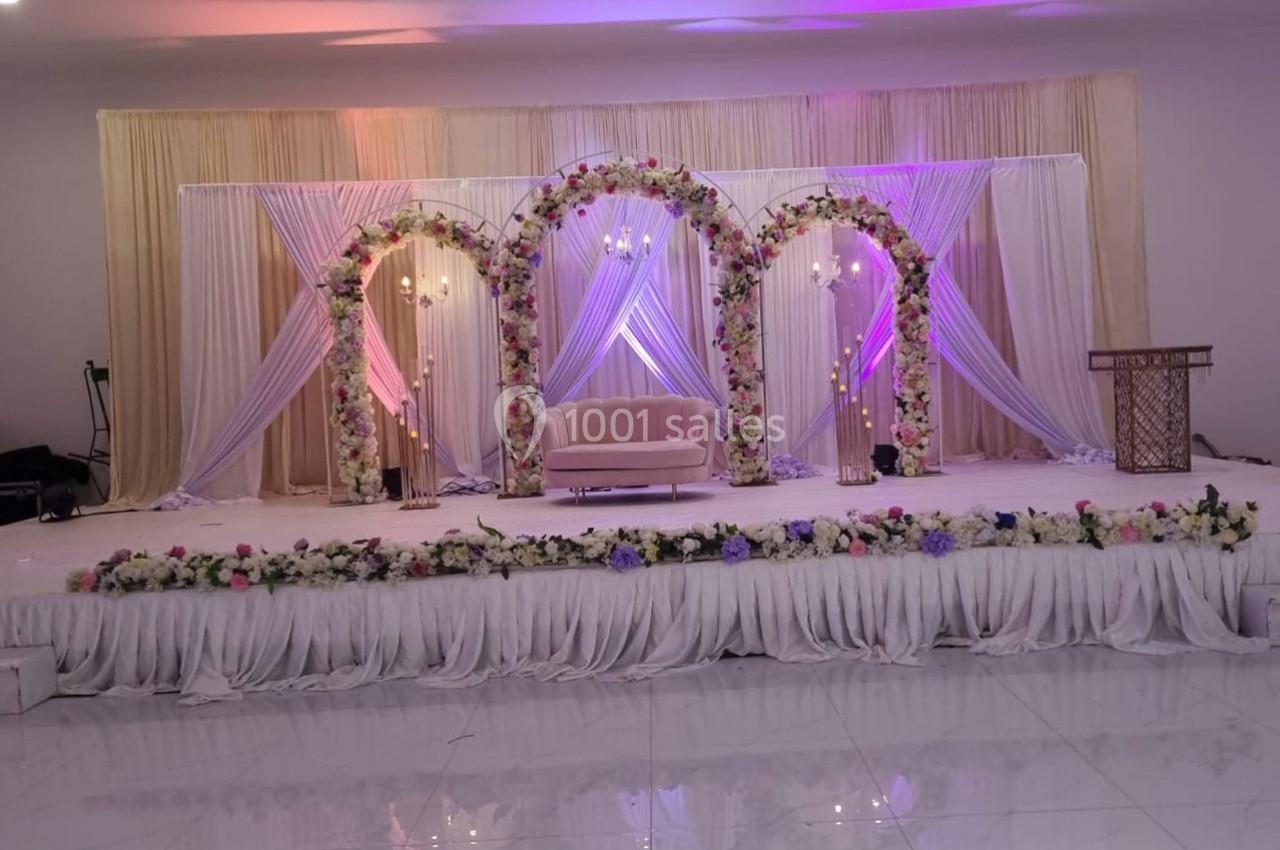 Décor de scène avec arche fleurie, rideaux lumineux, canapé rose et pupitre en bois sur fond de sol brillant.