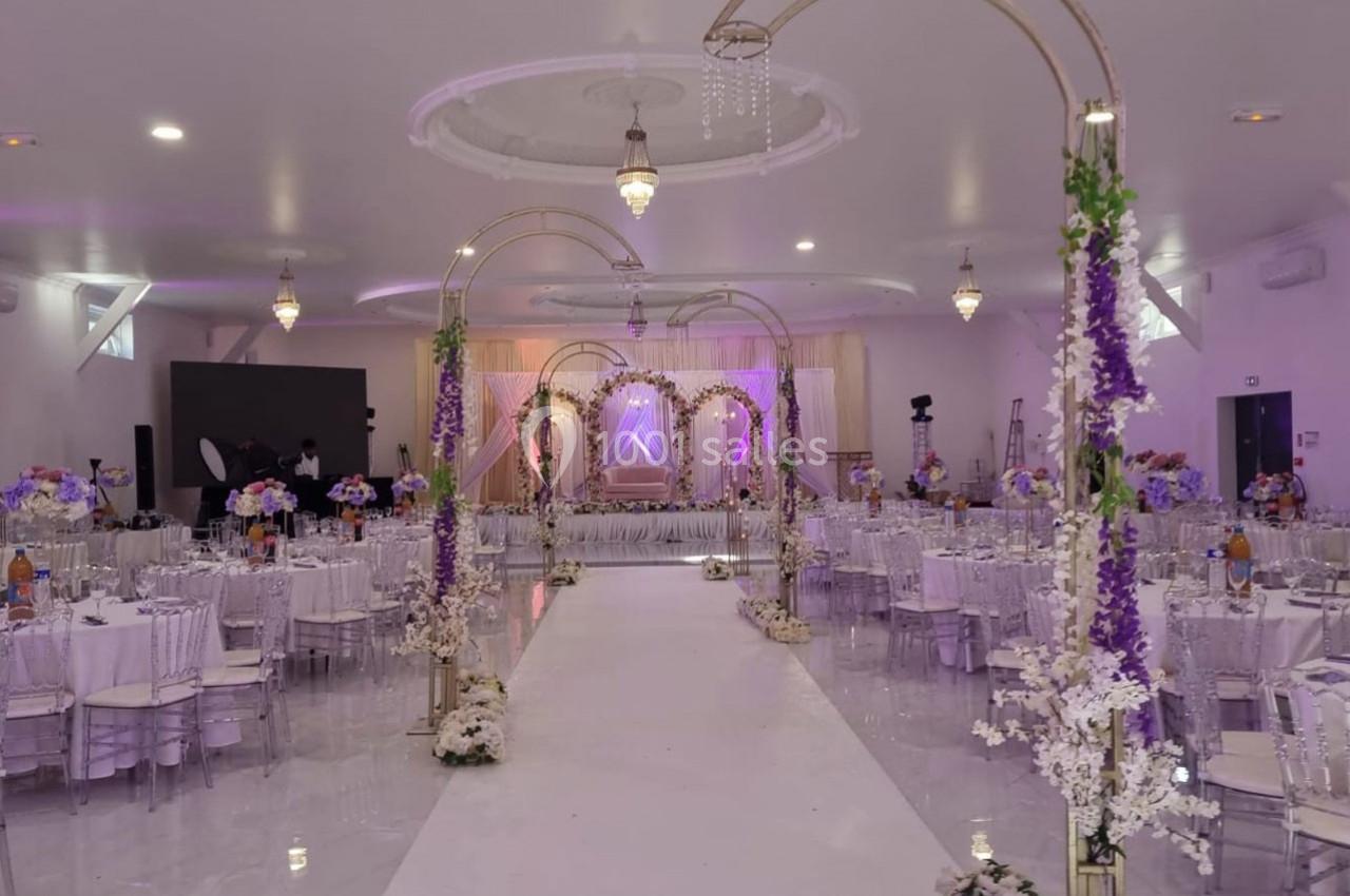 Salle de réception décorée avec des arches fleuries, tables dressées et éclairage tamisé pour un événement festif.