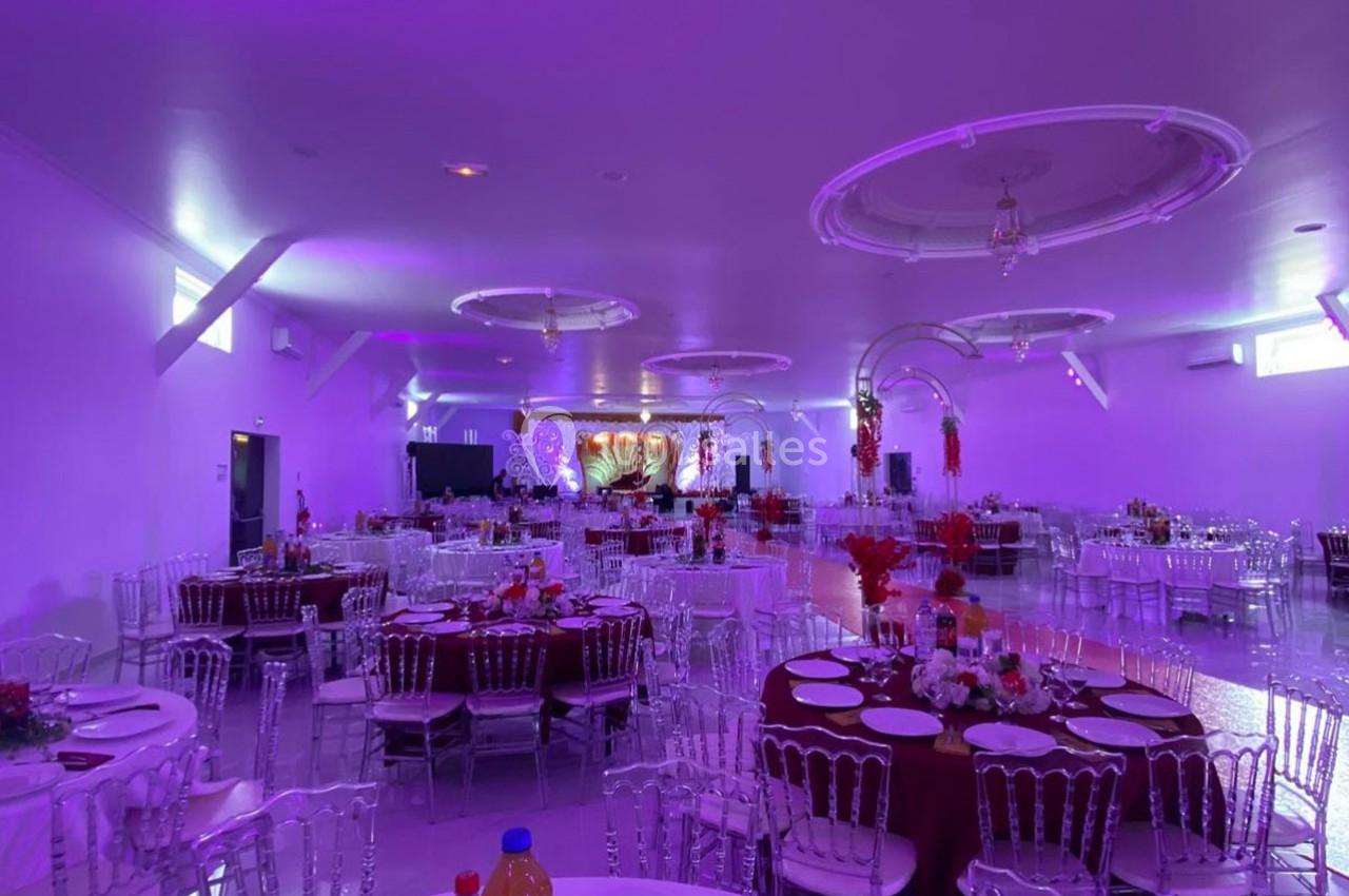 Salle de réception décorée avec des tables rondes dressées, éclairage violet et espace pour un événement festif.