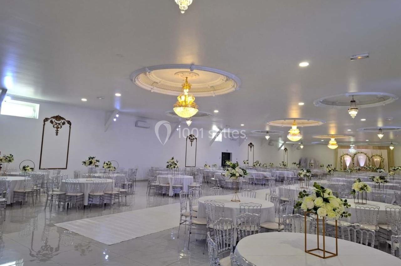 Salle de réception lumineuse avec tables rondes décorées de fleurs blanches et lustres suspendus au plafond.