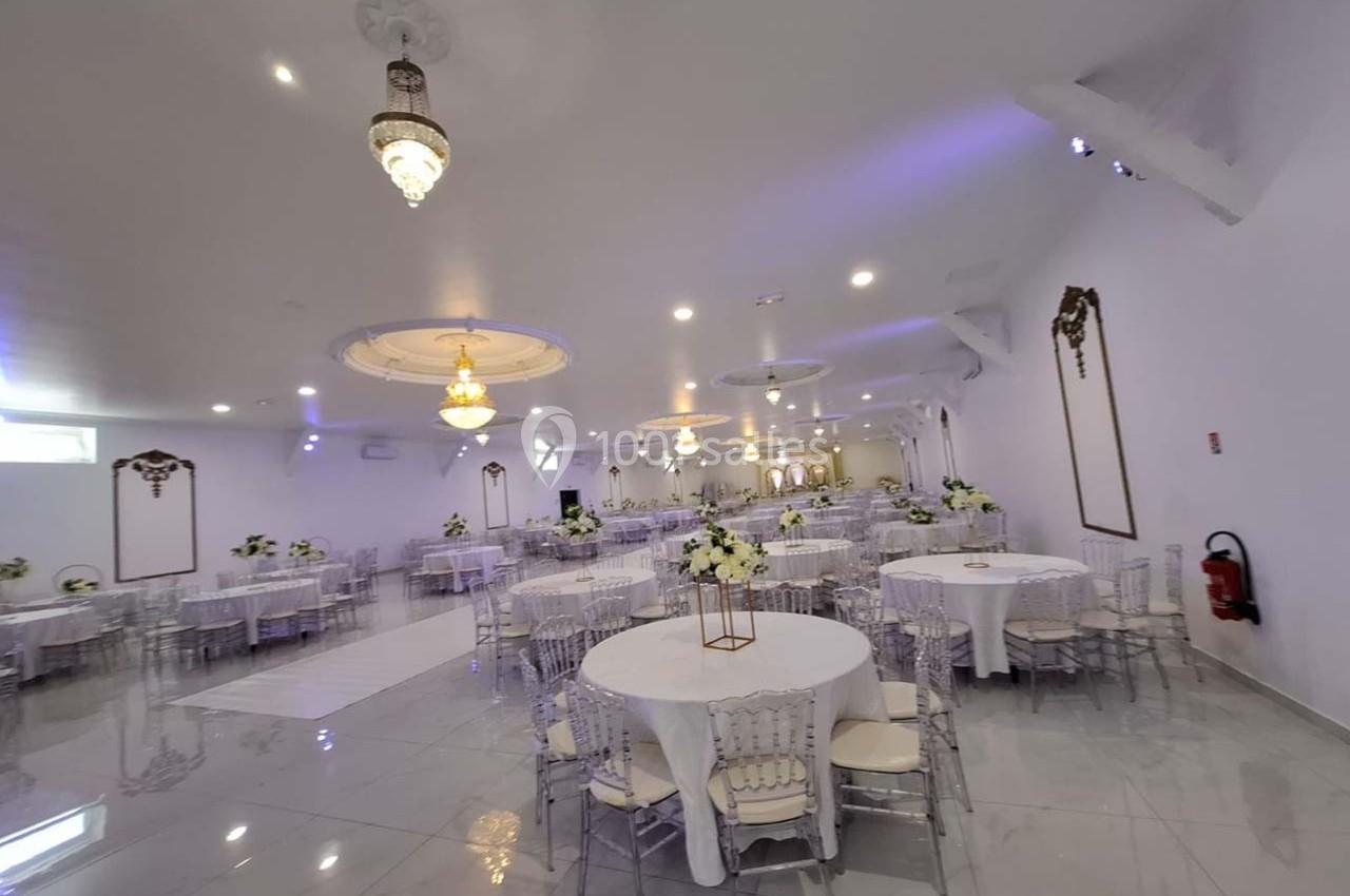 Salle de réception lumineuse avec tables rondes décorées de fleurs blanches, chaises transparentes et lustres élégants.