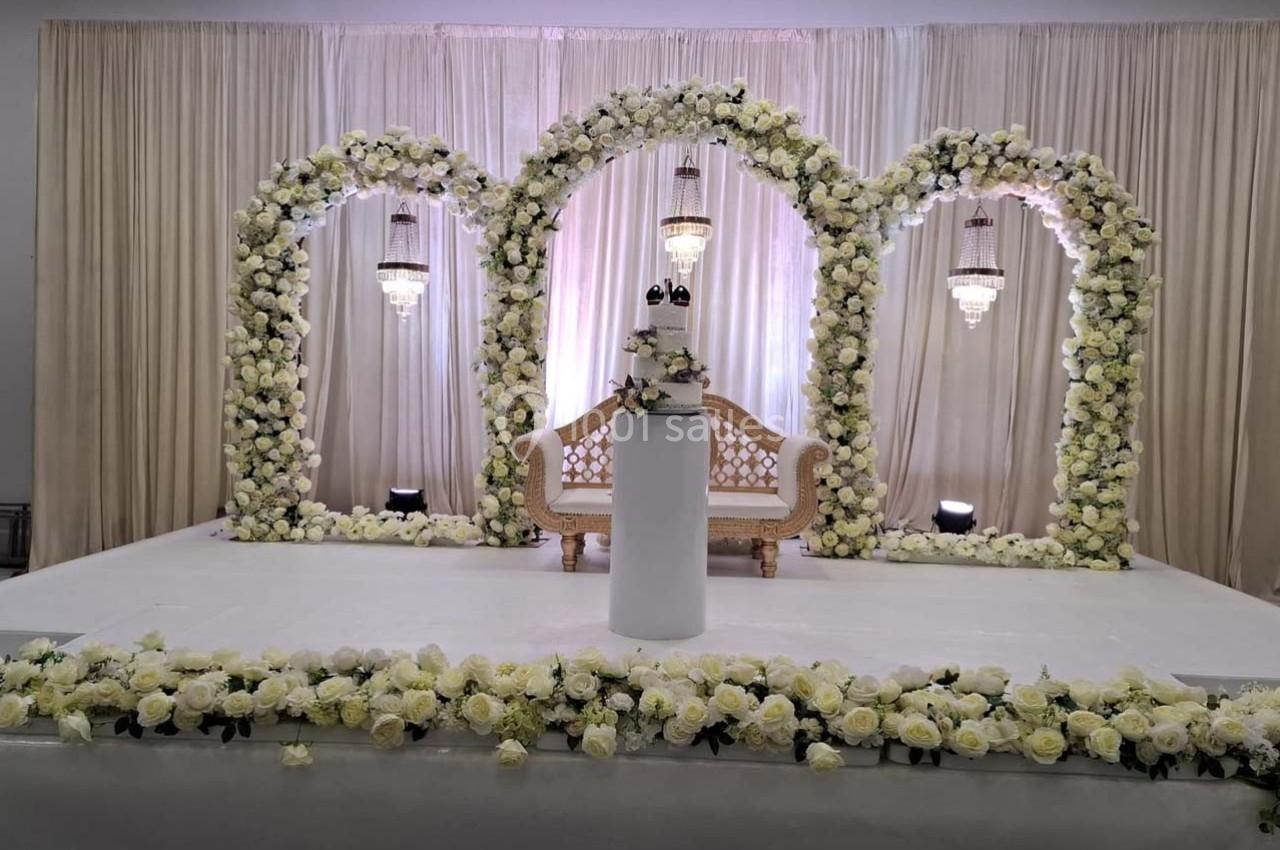 Décor de cérémonie avec arche florale blanche, canapé élégant et éclairage suspendu sur une scène ornée de fleurs.