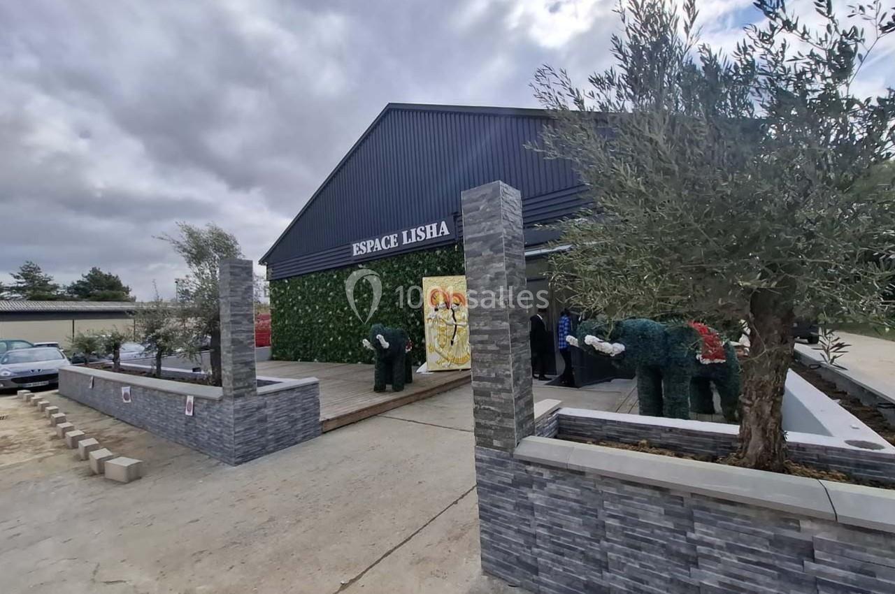 Bâtiment moderne avec façade noire, terrasse aménagée, murs végétalisés et sculptures, sous un ciel nuageux.