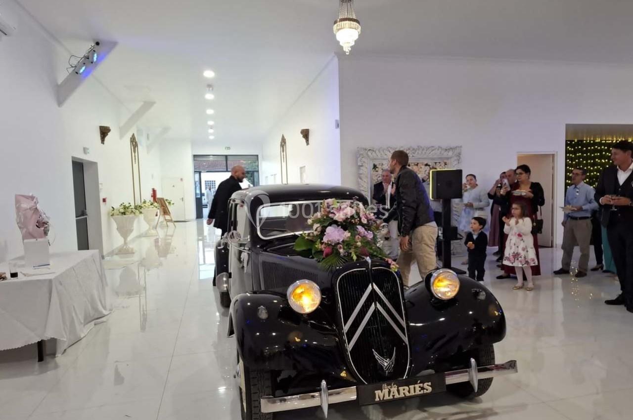 Voiture ancienne noire décorée de fleurs avec une plaque ’Mariés’, entourée d'invités dans une salle lumineuse.