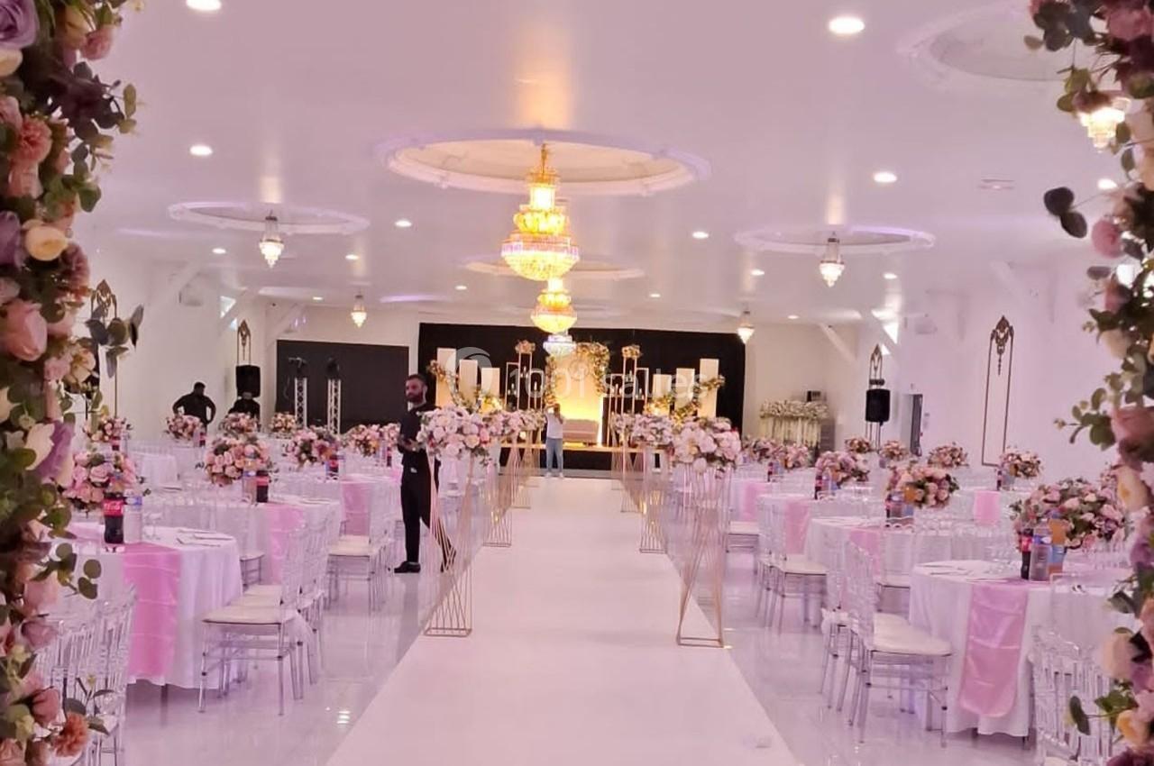 Salle de réception décorée avec des tables rondes, nappes roses, chandeliers dorés et allée centrale fleurie.