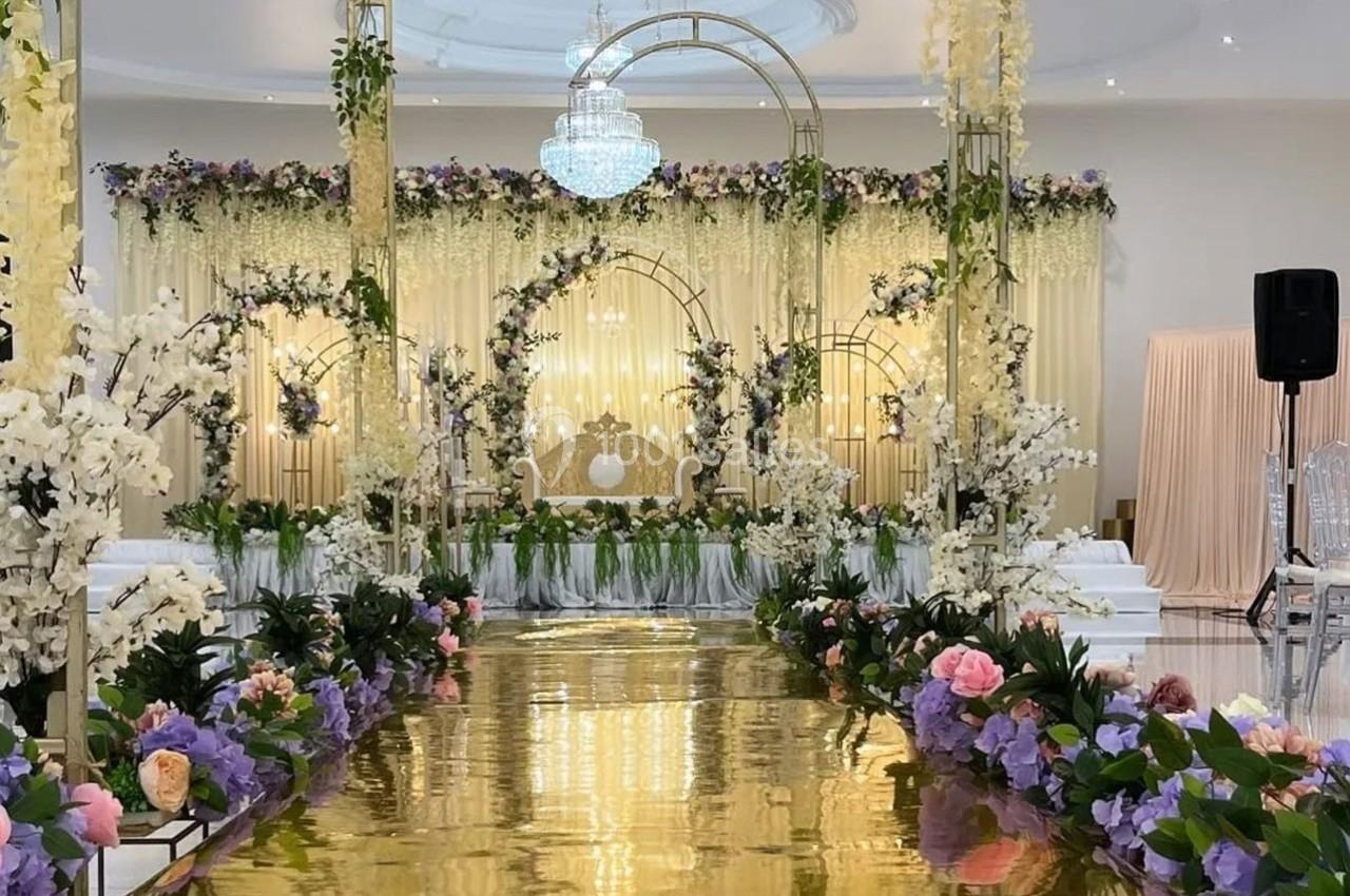 Allée dorée bordée de fleurs colorées menant à une arche décorée de guirlandes et d'un fauteuil dans une salle élégante.