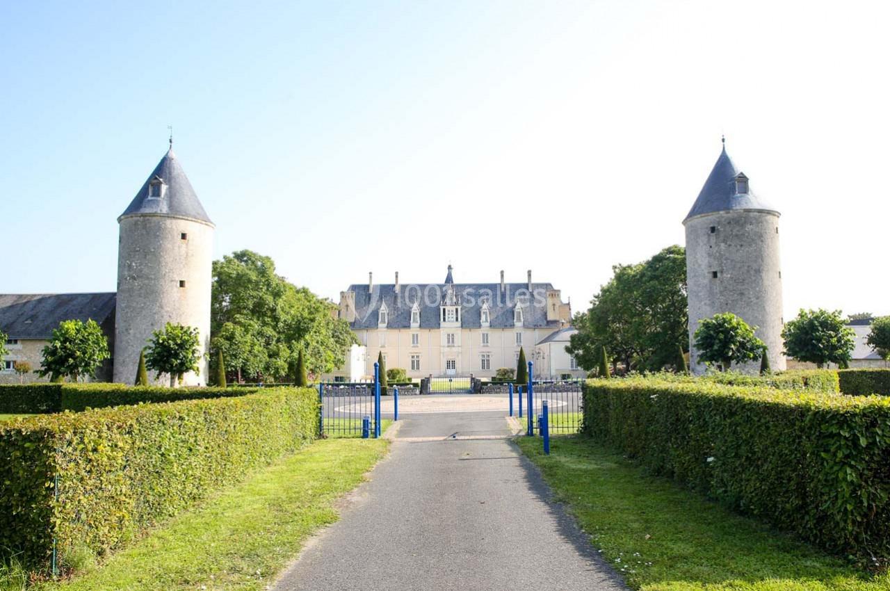Allée bordée de haies menant à un château avec deux tours rondes en pierre et un portail bleu.