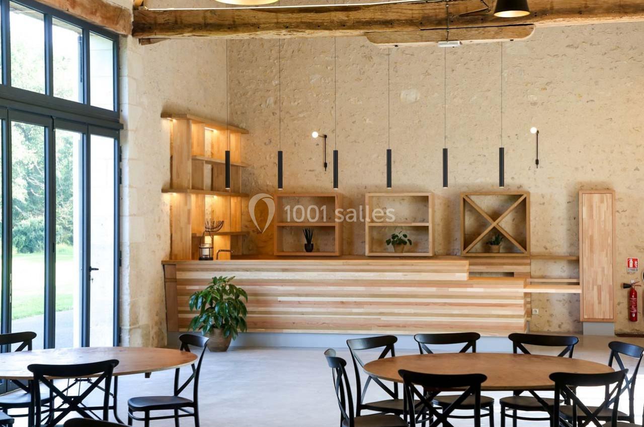 Salle lumineuse avec comptoir en bois, étagères ouvertes, tables rondes et chaises noires, murs en pierre apparente.