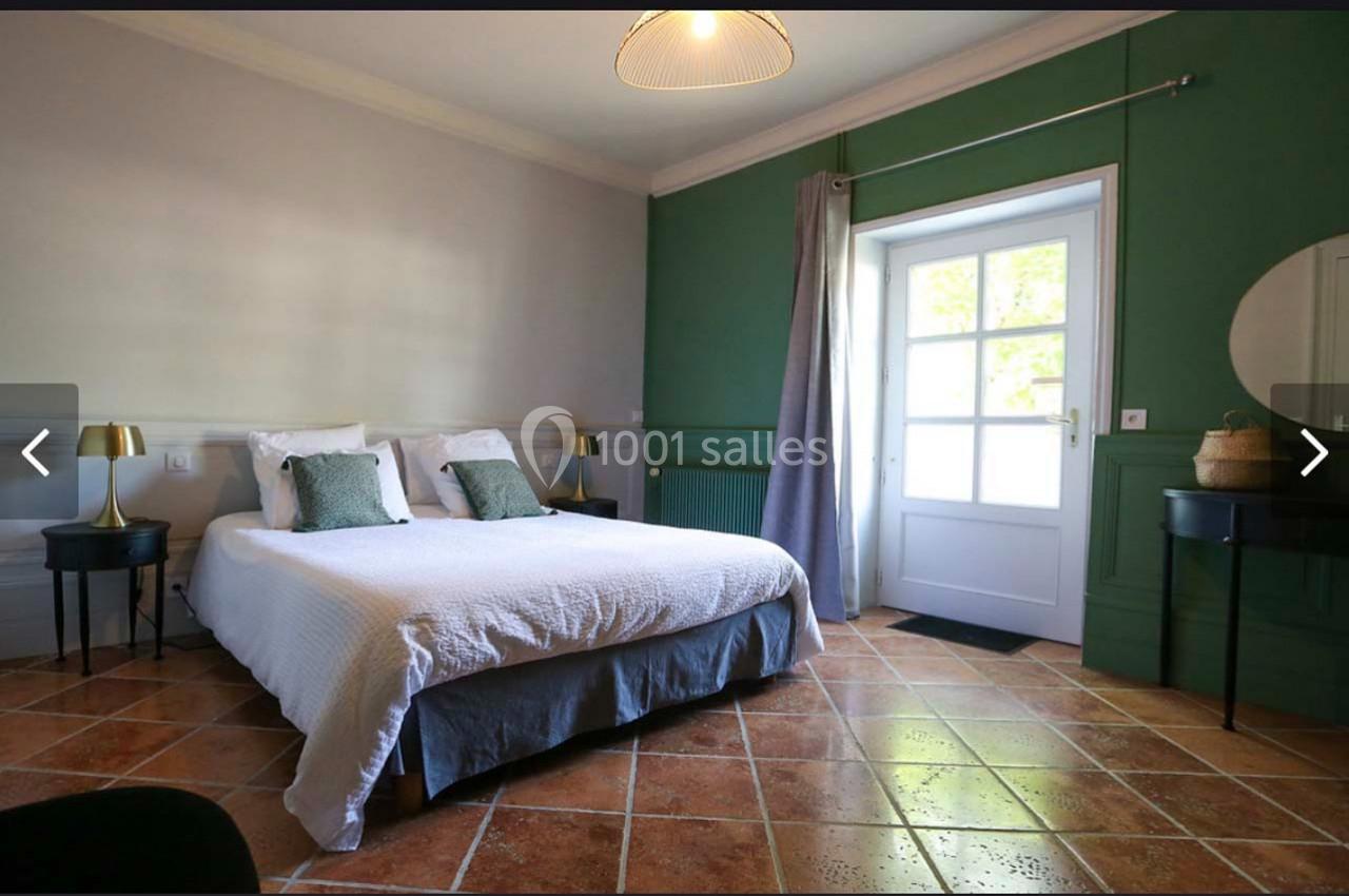 Chambre lumineuse avec lit double, murs verts et blancs, sol en carrelage, lampe suspendue et fenêtre donnant sur l…