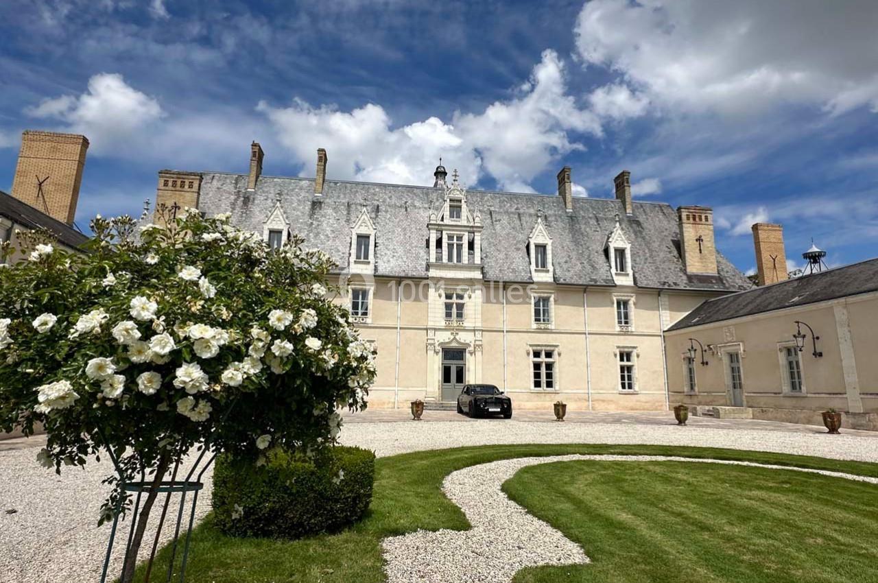 Façade d'un manoir en pierre avec toit en ardoise, jardin aménagé au premier plan et ciel partiellement nuageux.