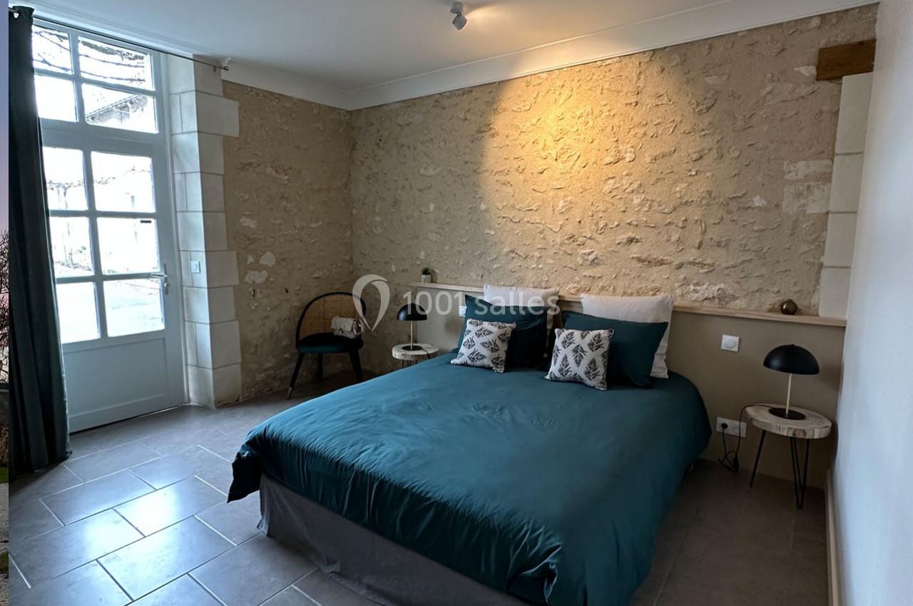 Chambre avec murs en pierre apparente, lit double avec draps verts, deux tables de chevet et une grande fenêtre.