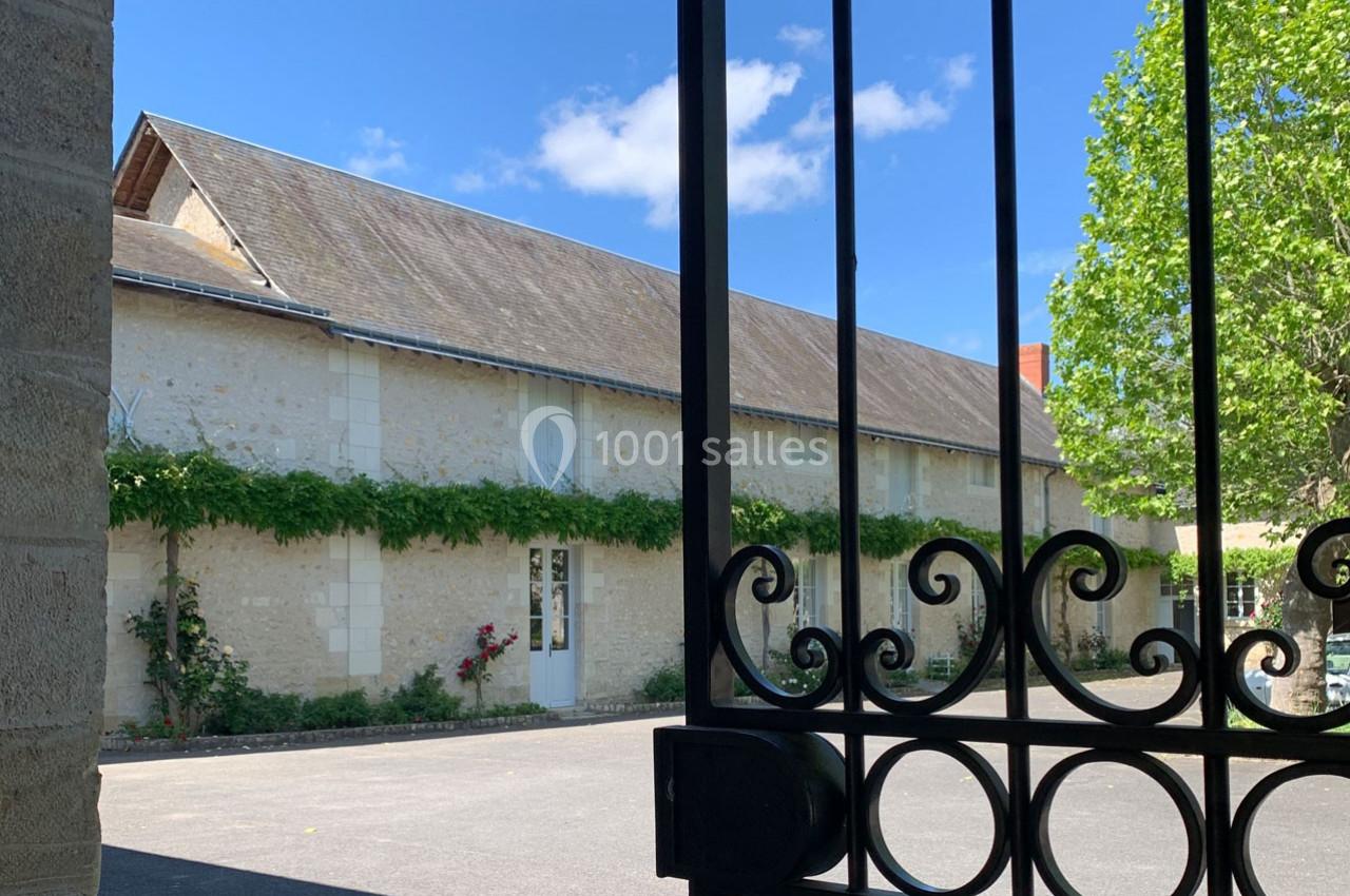 Bâtiment en pierre clair avec toit en pente, entouré de verdure, vu à travers une grille en fer forgé.