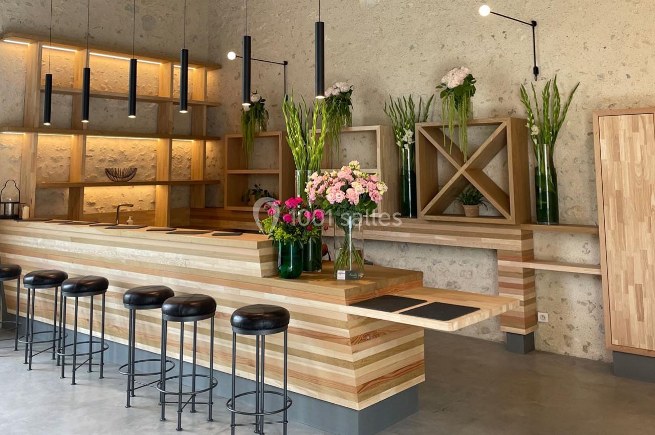 Comptoir en bois avec tabourets noirs, étagères décorées de fleurs et éclairage suspendu dans un espace moderne.