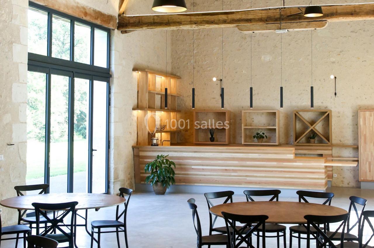 Salle lumineuse avec murs en pierre, grandes baies vitrées, tables en bois, chaises noires et comptoir en bois clair.