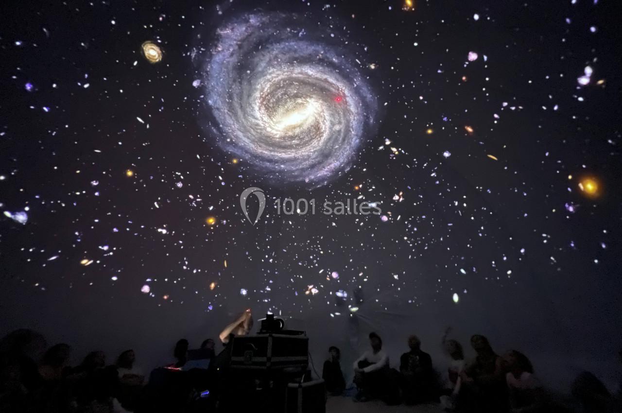 Projection immersive d'une galaxie spirale et d'étoiles dans une salle sombre, avec des spectateurs assis au sol.