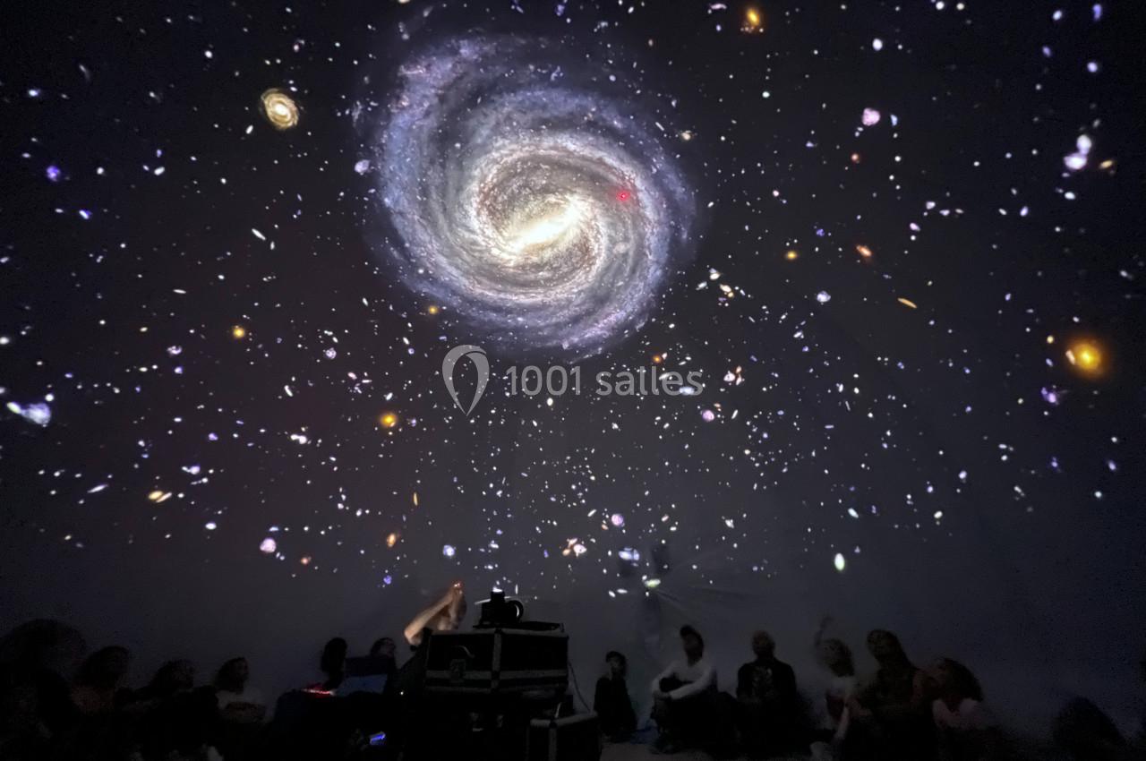 Projection immersive d'une galaxie spirale et d'étoiles dans une salle sombre avec des spectateurs assis au sol.