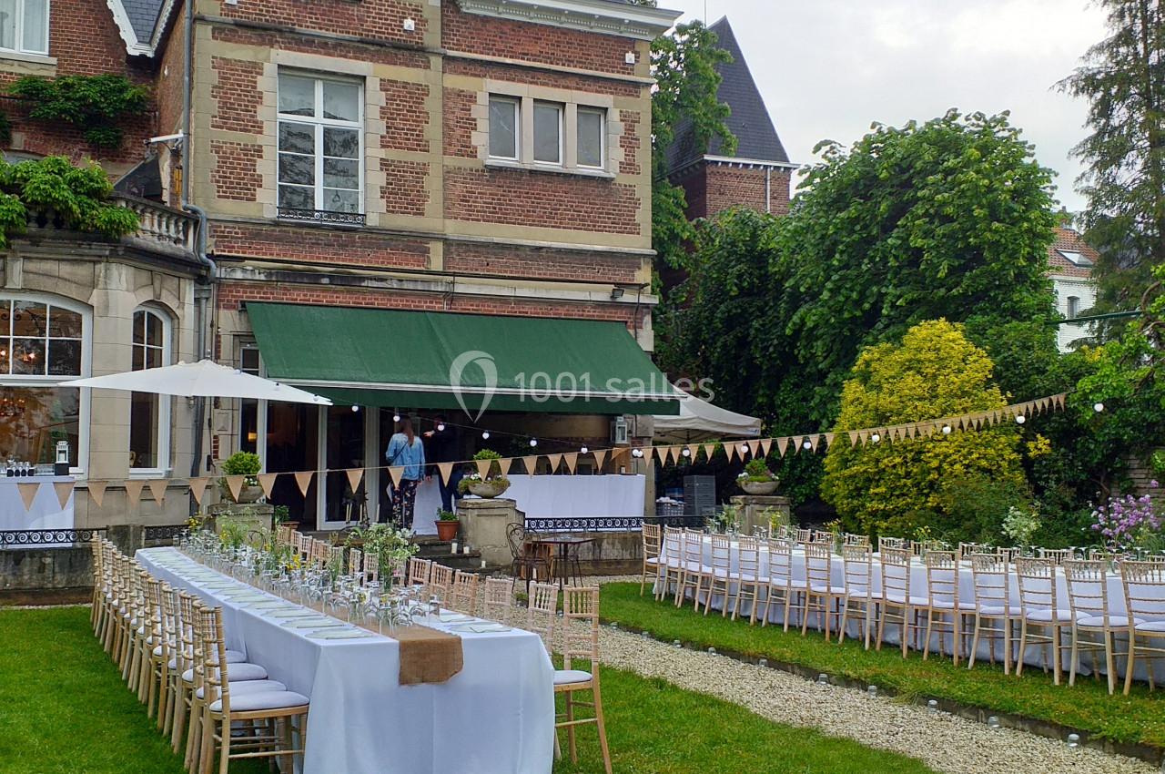 Tables dressées pour un événement en plein air dans un jardin, devant une maison en briques avec des guirlandes décoratives.