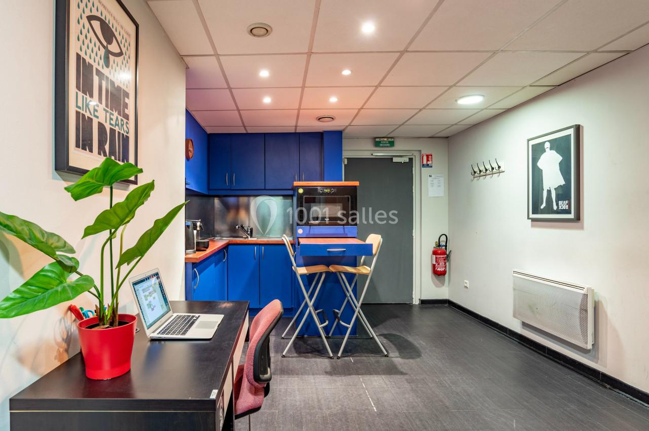 Coin cuisine avec meubles bleus, table haute et chaises, bureau avec ordinateur, plante en pot et décoration murale.