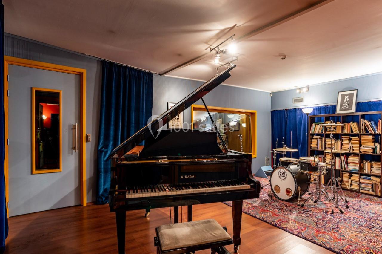 Salle de musique avec un piano à queue, une batterie, des étagères remplies de livres et un tapis coloré.